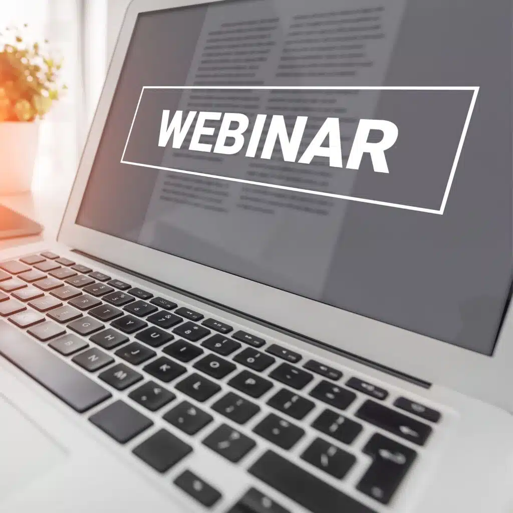 A laptop displaying the word WEBINAR in bold white text on the screen, with a blurred document and a potted plant visible in the background.