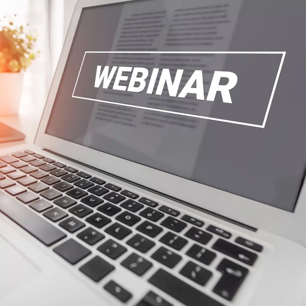 A laptop displaying the word WEBINAR in bold white text on the screen, with a blurred document and a potted plant visible in the background.