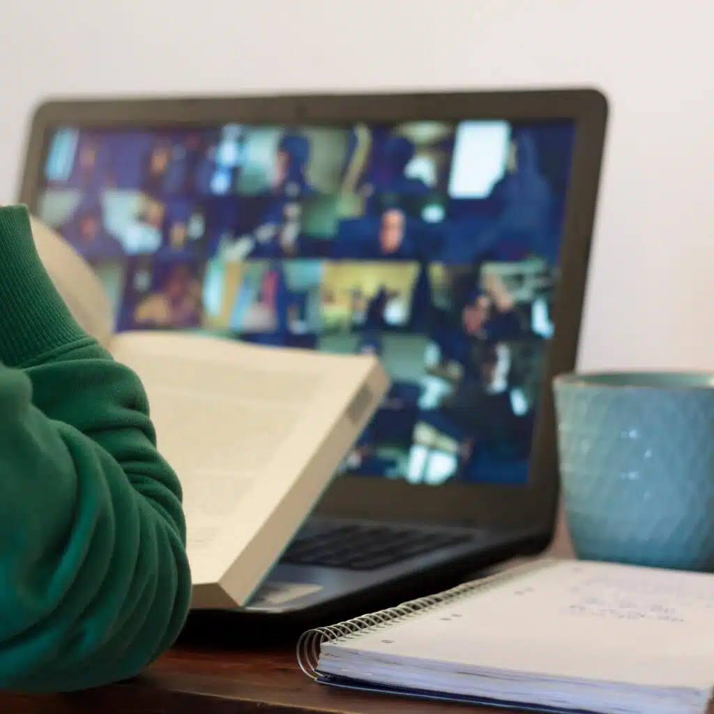 A person reads a book in front of a laptop showing a video call with many participants. A notebook and a blue mug are on the desk beside them.